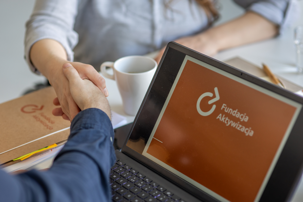 Zdjęcie przedstawia dwie dłonie w uścisku i komputer z logo Fundacji Aktywizacja.