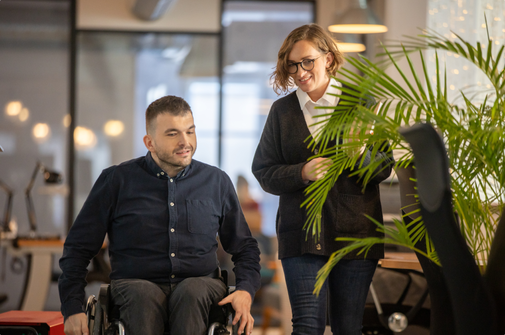 Mężczyzna poruszający się na wózku idzie korytarzem z kobietą. Rozmawiają i oboje lekko się uśmiechają.