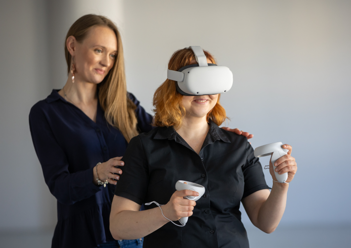 Na pierwszym planie kobieta ma założone gogle do VR i w dłoniach trzyma kontrolery. Z tyłu druga kobieta asekuruje ją, lekko trzymając ją za plecy. Obie są uśmiechnięte.