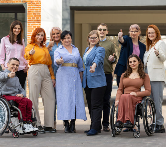 Grupa osób uśmiechniętych z kciukami do góry. Są to pracownicy i pracowniczki fundacji i klienci i klientki w tym dwie osoby na wózku, jedna o kuli i 3 słabowidzące.